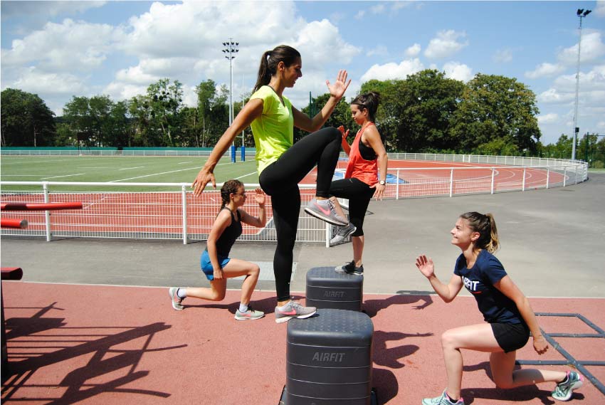 Four female athletes do training lunges using black step-up boxes by an outdoor track.